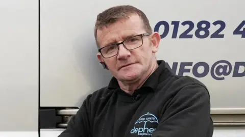 'Hero' plumber James Anderson's firm breached multiple rules, report finds Reuters James Anderson standing in front of his van. He is wearing a black jumper with branding for his company Defer. He has short brown hair and black glasses.