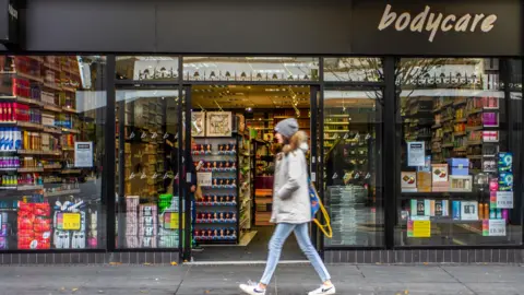 Bodycare to shut a further 30 stores after beauty chain collapse Alamy A person in a grey jacket and blue jeans walks past a shop with 'Bodycare' written on the top-right of the frontage. The store has large glass windows displaying various products such as toiletries, cosmetics, and household items. Shelves of products are visible inside the entrance. Signs on the windows advertise prices and promotions.