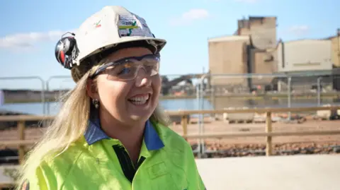 A blonde-haired woman wearing a hard hat, safety glasses and a high-visibility jacket stands at an industrial site. Behind her are buildings and fencing. It's a sunny day, with a few clouds in the sky.