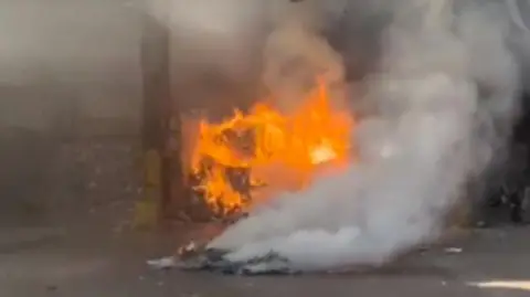 Vape ban isn't working, says waste firm boss Biffa A fire rages in a recycling facility. Plumes of smoke rise in the foreground, in front of a pile of material burning with bright orange flames.