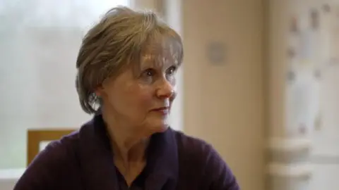 'Hero' plumber James Anderson's firm breached multiple rules, report finds Andrea looks to the side as she is interviewed by the BBC. She is wearing a navy blue blouse and has short light brown hair. A window to the countryside can be seen in the background.