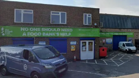 UK's first foodbank in Wiltshire celebrates 25th birthday Google A view of a single-storey small warehouse in Salisbury, red brick. On it are big signs in green for the foodbank with the Trussell Trust logo. It says "No one should need a food bank's help".