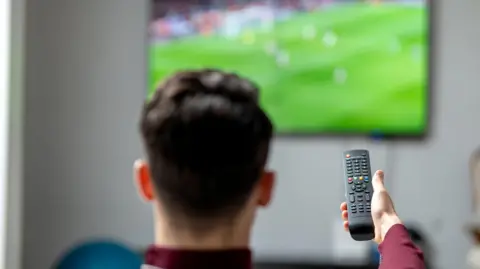 Largest illegal live sports streaming site shut down Getty Images A man sitting on a sofa watching a football match on TV, holding out a remote in front of him.