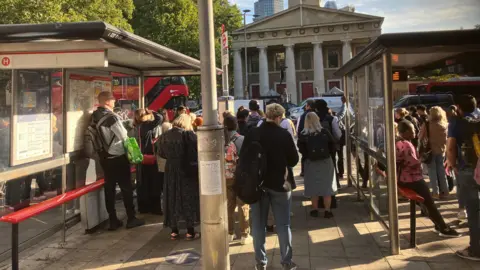 Londoners without Underground service during walkout A large group of people stand at a bus stop in central London on a sunny Monday morning. They have their backs to the camera.