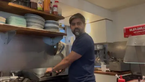 What impact will the Tube strike have on the economy? Prasanna Callaghan A middle aged man with a greying beard stands in a cafe kitchen. He is staring into the camera. There are many plates and condiment bottles on shelves in front of him.