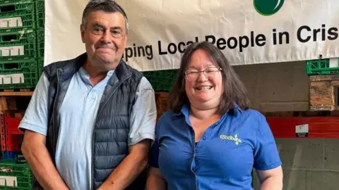 UK's first foodbank in Wiltshire celebrates 25th birthday BBC Mike and Maria smile at the camera while standing in a warehouse, part of a foodbank banner behind them. Mike is wearing a blue polo top and dark gilet while Maria wears a blue shirt with foodbank branding.