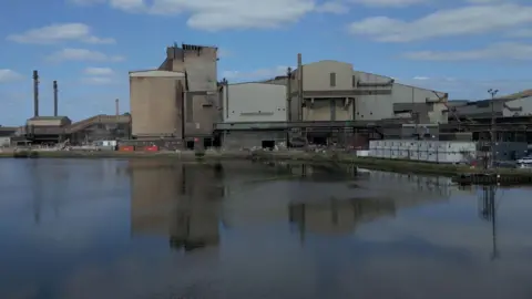 Tata Steel UK An industrial steelmaking site in Port Talbot, South Wales, seen from across a calm body of water. Several large buildings dominate the scene, including tall chimneys and metal-clad structures. Construction materials and equipment are visible near the buildings, suggesting ongoing work. The buildings are reflected clearly in the water, and the sky above is partly cloudy with patches of blue.