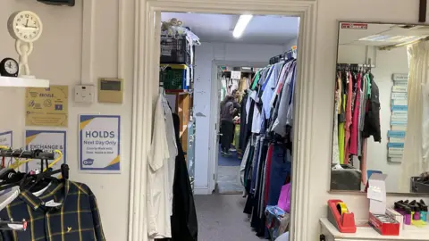 Spike in charity donations from people leaving Jersey Indoor shot of the entrance to a charity shop clothing storeroom. In the foreground, on the left, a rack of hanging clothes holds a dark plaid button-up shirt. Next to it, a blue sign with yellow text reads "HOLDS Next Day Only" with a list of names underneath. A doorway in the center leads to a smaller room with more clothes hanging on racks. In the distance, a person is partially visible, walking away. On the right, a large mirror reflects the room and the clothing racks.