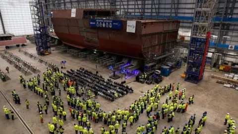 Rosyth shipyard expected to win £1bn Danish navy contract Getty Images Crowds of people in fluorescent yellow safety jackets and hard hats viewed from above. They are in a large warehouse looking at a section of a brown warship in construction.