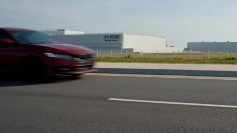 Getty Images A red car drives by the Hyundai plant in Georgia, a large white building behind a fence in a grassy field. 