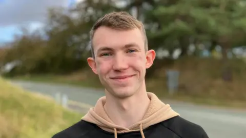 University students like me are happier living at home Isaac, a young man wearing a beige hoodie with a black jumper, with short dark blonde hair, smiles at the camera
