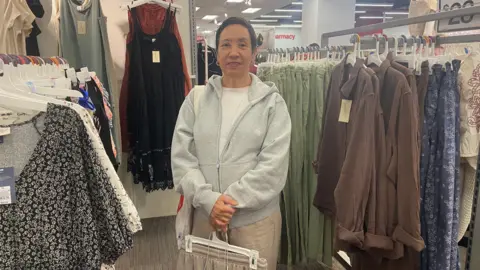 Rising US prices could widen divide between haves and have-nots Danielle Kaye/BBC Nancy Garcia, who works in the publishing and gifts industry, shops for clothing at a Target location in Manhattan on 10 Sept 2025. She is surrounded by dresses and jackets on hangers and is holding some clothes.
