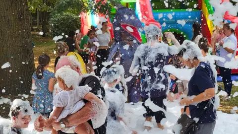 Cambridgeshire Send charity 'supported 8,000 people' in 'August Little Miracles Children and parents playing with white foam- with a colourful bouncy castle in the background.