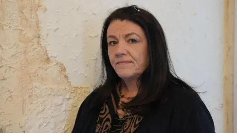 30,000 homes fitted with botched insulation under government schemes, ministers admit Susan Haslam with long dark hair and a black cardigan and brown and black patterned blouse looks at the camera with a negative expression as she stands in front of a white wall with a huge patch of brown damp