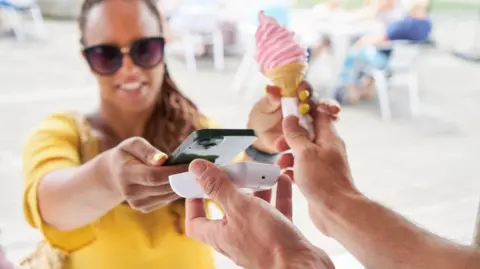 Getty Images A woman dressed in a yellow top and wearing sunglasses buys a pink ice cream by placing her smartphone near a payment terminal.