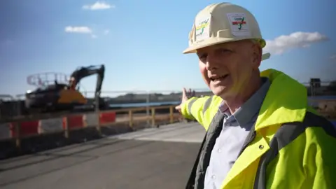 A man in a white hard hat and high-visibility jacket stands on a construction site, pointing toward an excavator in the background. Safety barriers line the area, and the sky is partly cloudy.