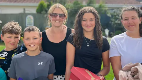 'Less of a stigma' on second-hand school uniforms BBC (left to right James (10) , Henry (11), Mum Nikki, Grace (14) and Olivia (16)). They are all smiling holding their back to school purchases and hand-me downs. The family is standing in the garden on a sunny day. They all have black hair apart from the mum who is blonde.