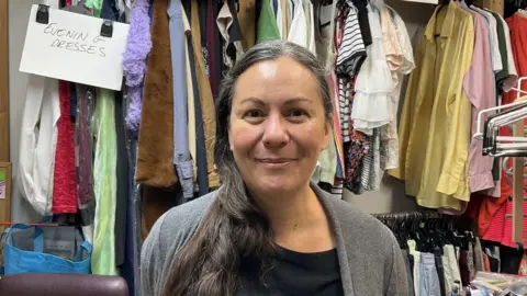 Spike in charity donations from people leaving Jersey BBC Elk Hargreaves stands in a charity shop, with racks and racks of clothes behind her. She has long dark hair in a ponytail over the shoulder and is smiling.