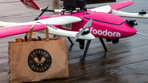 Will drones deliver your next hot food order? Aviant The foodora drone is pink and white with rotors on mounts attached to the wings - a third propeller is on the back. In the foreground is a brown paper bag from Bastard Burgers.
