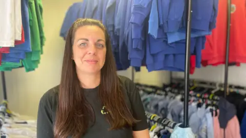 School costs a 'real struggle' for parents in Wales A woman with long brown hair, wearing a black T-shirt is standing in front of rails of uniform arranged by colour