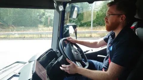 Lack of charging facilities a 'barrier' for electric lorries BBC Lorry driver Liam Ely sitting at the wheel as he drives down a road