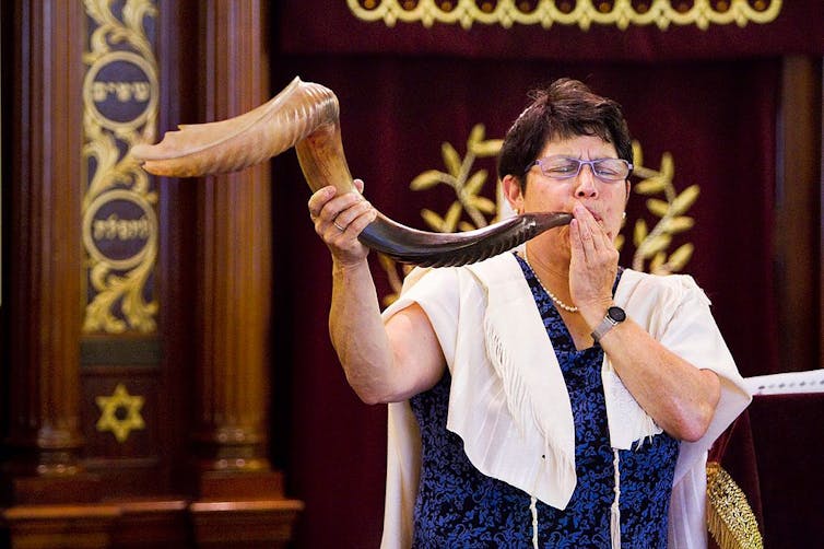How the spiritual sound of the shofar shapes the Jewish new year – a Jewish studies scholar explains A woman in a blue dress and white prayer shawl blows into a large animal horn.