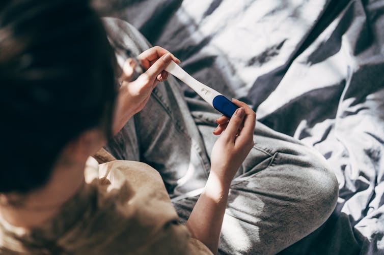 A brunette woman, her face unseen, sits on a bed as she holds a pregnancy test in two hands.
