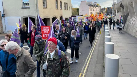 A large march taking place along The Royal Mile. Participants are carrying purple flags with “Unison” branding and banners with messages such as “Scotland Demands Better – The Mandate From All of Us.” In the background, more demonstrators hold bright red and yellow flags, creating a colourful and organized protest scene.