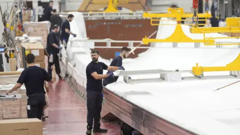 Government vows to create 400,000 jobs in clean energy sector PA Media A spacious industrial workshop where several workers in dark clothing are assembling or inspecting large white objects. Yellow overhead cranes and lifting equipment are visible above. The workspace is clean and organized, with various tools and machinery around.