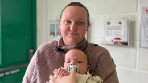 BBC Bethany Walker wearing a mauve knitted jumper with a black microphone clipped to the collar. She has brown hair pulled back in a bun and is holding her three-mont-old baby, who has a blue pacifier in his mouth.