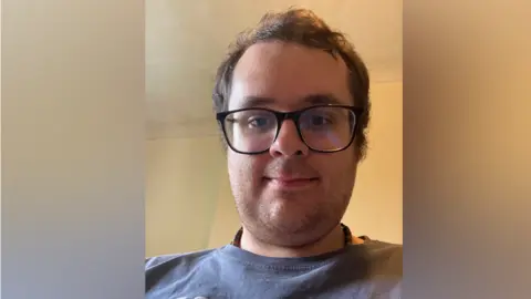 What is driving the decision to learn in a manual or automatic car? David Robinson David, a white man with brown hair and stubble. He is smiling slightly at the camera and wears glasses