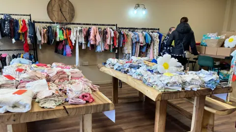 Baby and children's clothes are folded on tables and hang on racks. Two women are standing next to nappies. 