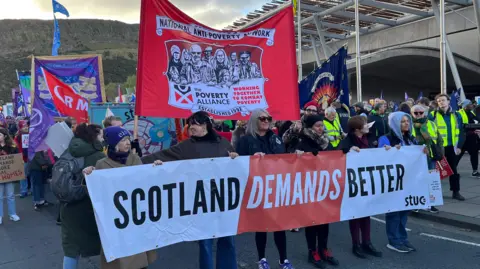 BBC A large protest march with participants holding banners and flags. The main banner in the foreground reads “SCOTLAND DEMANDS BETTER” and includes the STUC logo. Behind it, another prominent red banner displays “National Anti-Poverty Network” and “The Poverty Alliance – Working Together to Combat Poverty.” Several other colourful flags and signs are visible, and uniformed stewards stand along the side of the crowd.