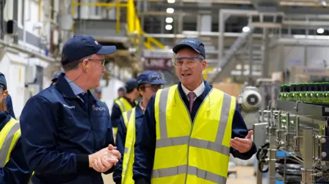 UK targets £420m at sky high industry energy bills PA Media Peter Kyle being led around a glass factor in Cheshire - he is gesturing with his left hand, while standing among the machinery - he is wearing a baseball cap and a high visibility jacket