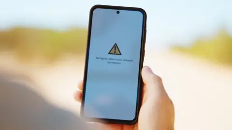 Getty Images A person holds a phone with a plain white background. On the screen is an error message stating there is no phone signal.