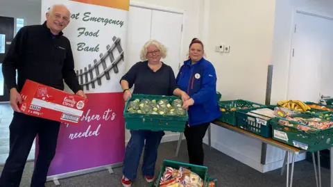Didcot Foodbank Three volunteers at the food bank show some of the donations. A man with grey hair and a moustache is holding a box of biscuits. A blonde woman with red-rimmed glasses and a woman with brown hair in a Tesco uniform hold a large crate of broccoli between them. More groceries are on the table behind them.