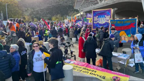 A busy outdoor gathering shows numerous people participating in a demonstration near the modern Holyrood building with hills in the background. The scene is filled with colourful banners and flags, including ones reading “STUC Youth Committee” and “Stand Up for Education.” 