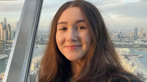 What is driving the decision to learn in a manual or automatic car? Cleo Moseley Cleo, a white woman with long brown hair. She is standing in front of a window with a city skyline in the background