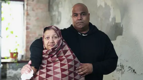 Government urged to review botched insulation schemes across Britain Bushra and Atif standing close together indoors against a wall with peeling paint and visible damage. Atif is wearing a dark hooded sweatshirt and has an arm around Bushra, who is wrapped in a patterned shawl with red and beige triangles. A window with a potted plant is partially visible in the background.