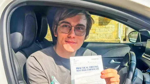 What is driving the decision to learn in a manual or automatic car? Luke Breaban-Cook Luke, an 18-year-old white man, sitting in the driver's seat of a car. The car door is open and he is facing outwards, holding a certificate showing he passed his driving test