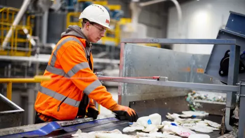 Can the plastic recycling industry be saved? Biffa A man in an orange hi-viz jacket stands at a conveyor belt carrying squashed plastic bottles.