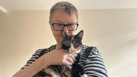 Pet owners say they are forced to cut back on food to afford vet bills Lucy stands holding her tortoise shell cat. The cat is black, brown and white with green eyes. The cat is looking at the camera. Lucy wears a black and white striped top with black overalls. The photo is against a cream wall.