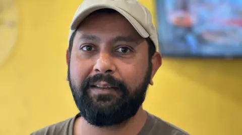 Workers back minimum wage rise 'as everything getting expensive' BBC Hassan Ali Tipu, who has a dark beard and it wearing a brown T-shirt and a cream coloured baseball cap.