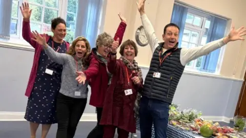 Positive Community Action celebrates 8,000 meals in Wiltshire Positive Community Action Five people, four women and one man, smile with their arms outstretched with food laid out on a table behind them.