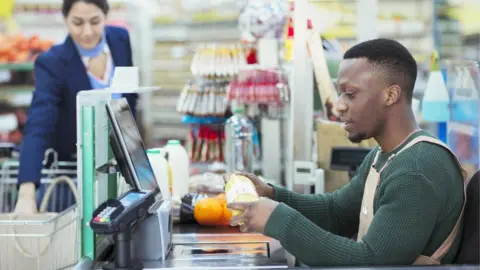 How much are the National Minimum Wage and National Living Wage worth? Getty Images A young man wearing a green sweater and a beige apron works on a supermarket checkout. He is serving a woman wearing a blue and pink top and navy jacket.