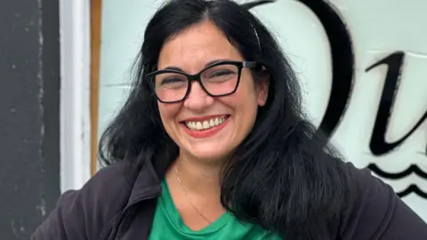 Workers back minimum wage rise 'as everything getting expensive' Matilde Liso, who has long dark hair and is wearing black rimmed glasses and a black top over a green T-shirt. She is smiling.