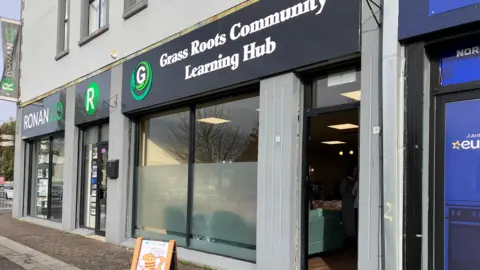 A building. The sign says Grass Roots Community Learning Hub. There's a board outside with an image of a bee. 
