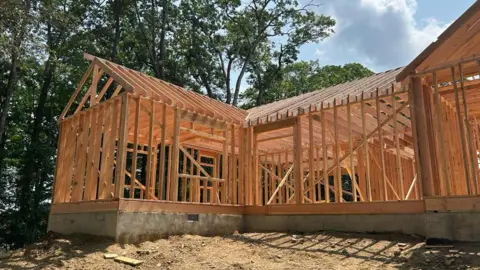 New Trump tariffs pose another hurdle for the housing market Anthony Cabrera A house under construction, with only the wooden frame built, on a dirt surface. next to trees against a blue cloudy sky.