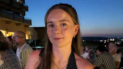 What is driving the decision to learn in a manual or automatic car? Caitlin Graham A close up photo of Caitlin, a young woman with long brown hair. She is smiling against a dusky night sky.