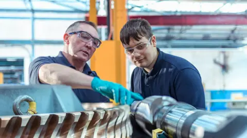 How much are the National Minimum Wage and National Living Wage worth? Getty Images An older man wearing blue plastic gloves is showing a large piece of metal equipment to a young man, both of whom are wearing short-sleeved navy polo shirts. They also have protective eye wear on and appear to be stood in a large factory warehouse, which is blurred in the background behind them.